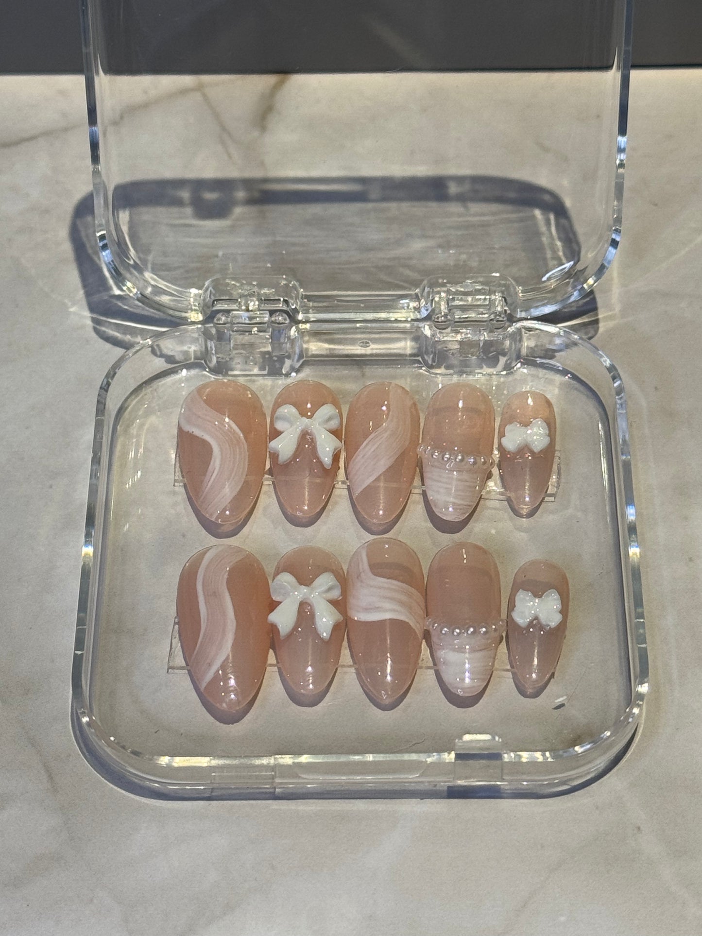 edium almond-shaped press-on nails in soft nude tones with delicate brushstroke detailing and charming bow and pearl embellishments. Each nail features hand-painted artistic strokes that add dimension and sophistication to your manicure. The combination of subtle nude shades with decorative accents creates a natural yet elegant look that complements any style, from everyday wear to special occasions.