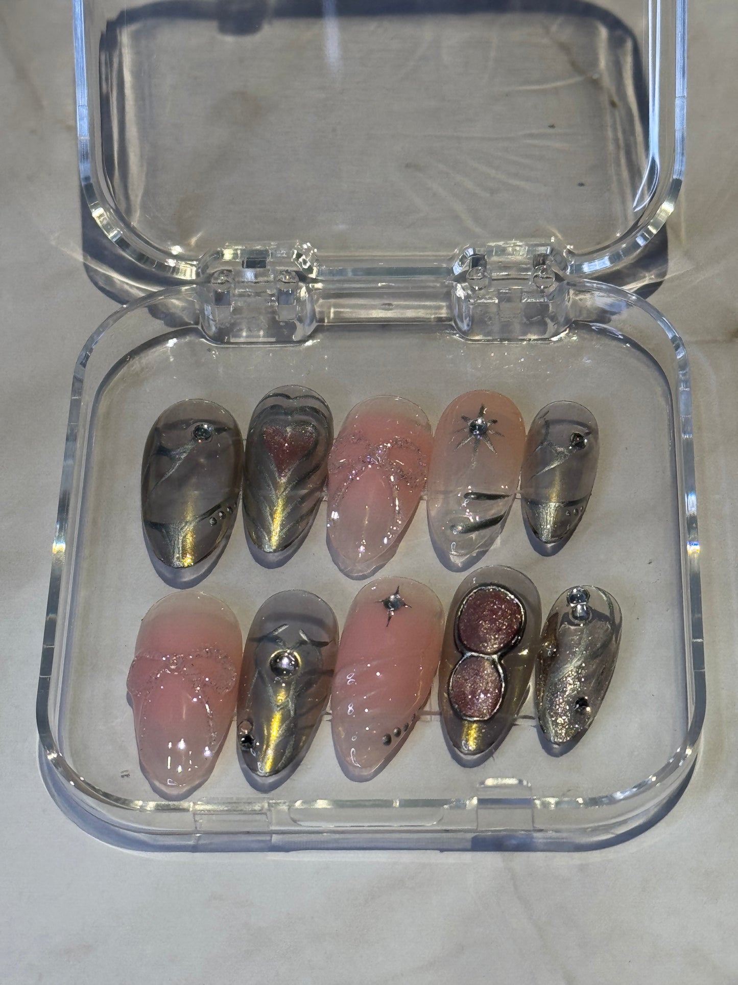 Medium almond shape with intricate details crafted for a bold, regal look. Features rhinestone embellishments, eye-detailed silver accents, and dark silver 3D effects for dimension. Soft pink ombre fades into gel details—all handmade for a unique warrior-inspired nail design that stands out. løsnegler